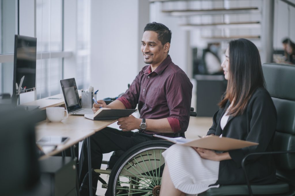 Indian white collar male worker in wheelchair having cheerful discussion conversation with his female asain chinese colleague coworking in creative office workstation beside window.