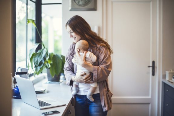Smiling mother using laptop while carrying baby at kitchen counter. Woman is working while holding son at home. She is wearing casuals.